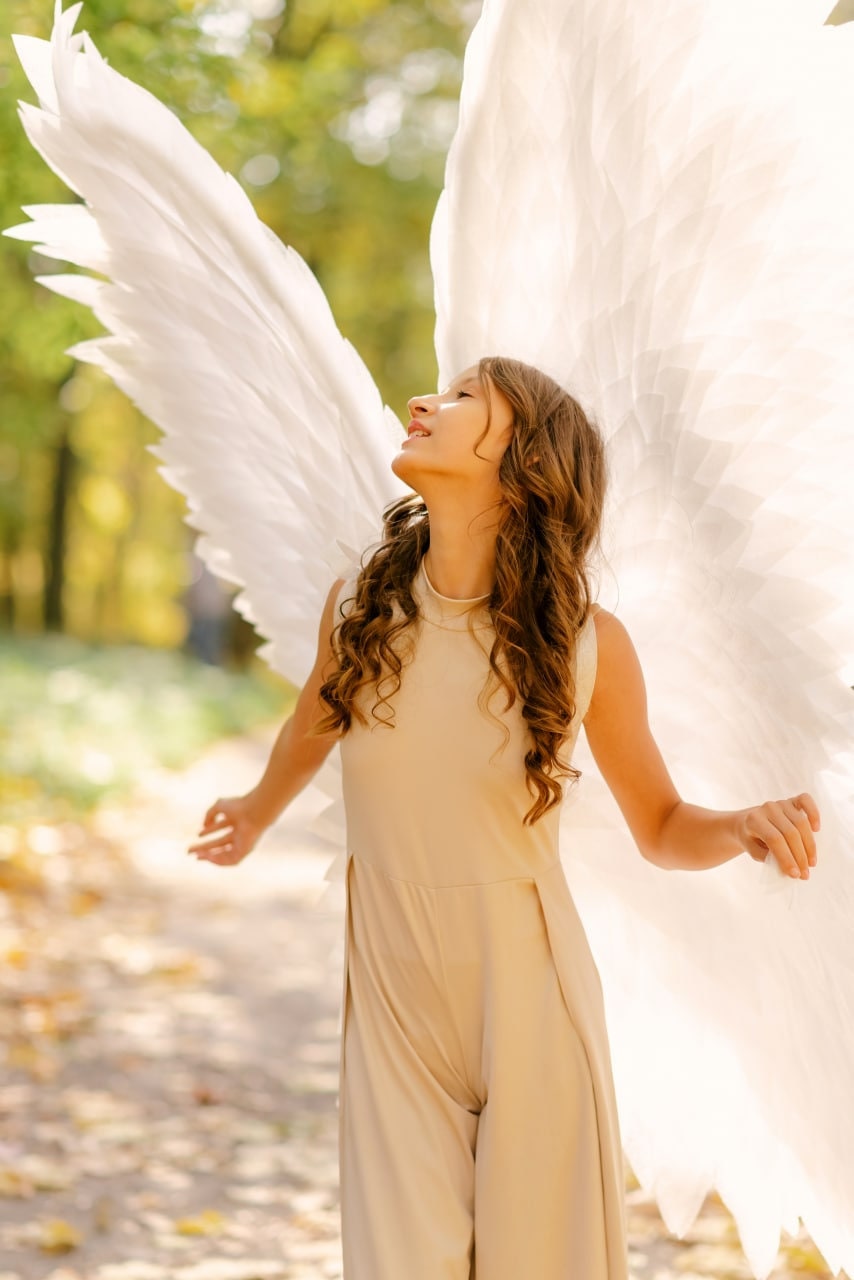 May include: A young person wearing a beige jumpsuit with white angel wings. The person is looking up and smiling. The background is a blurry image of a forest.