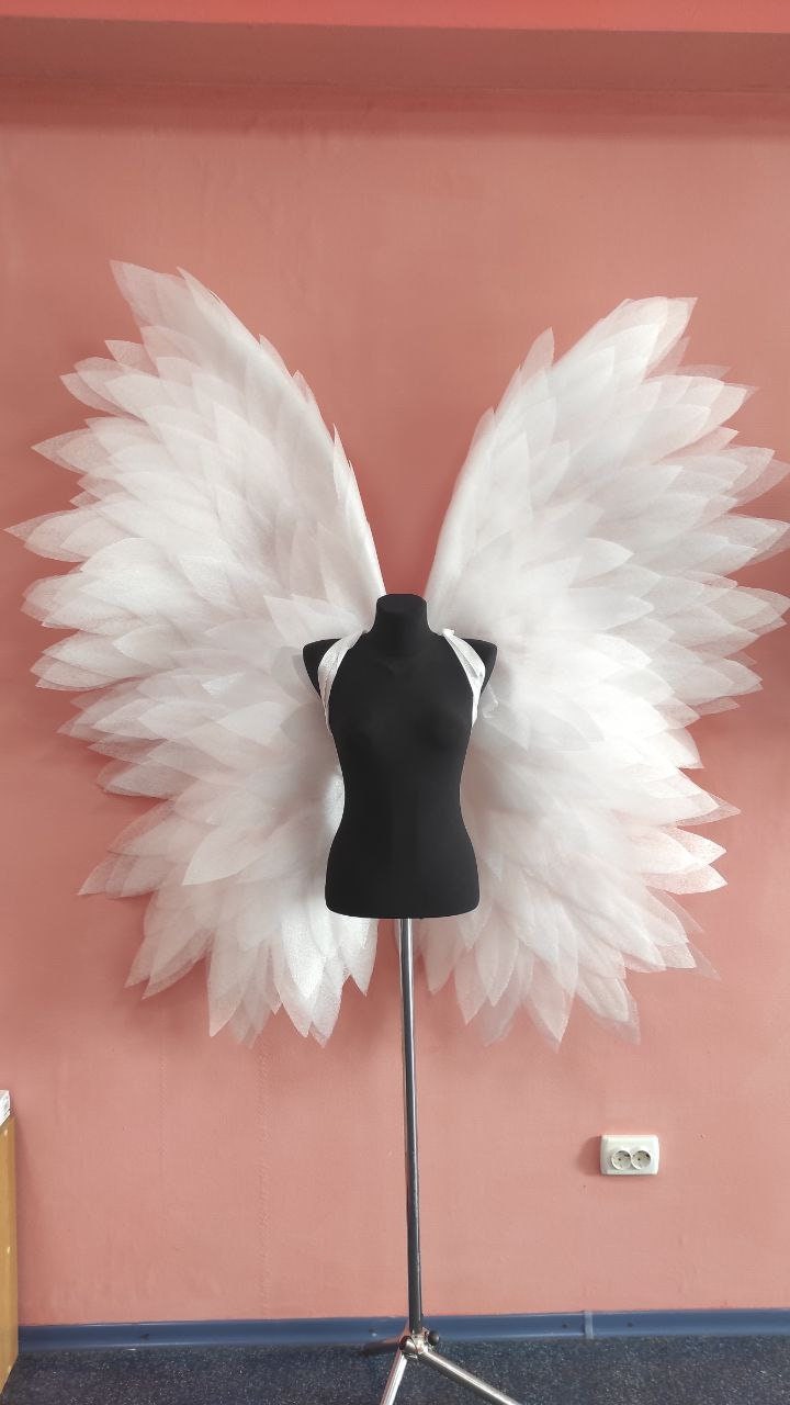 May include: White angel wings made of layered tulle fabric, displayed on a black mannequin stand against a pink wall.