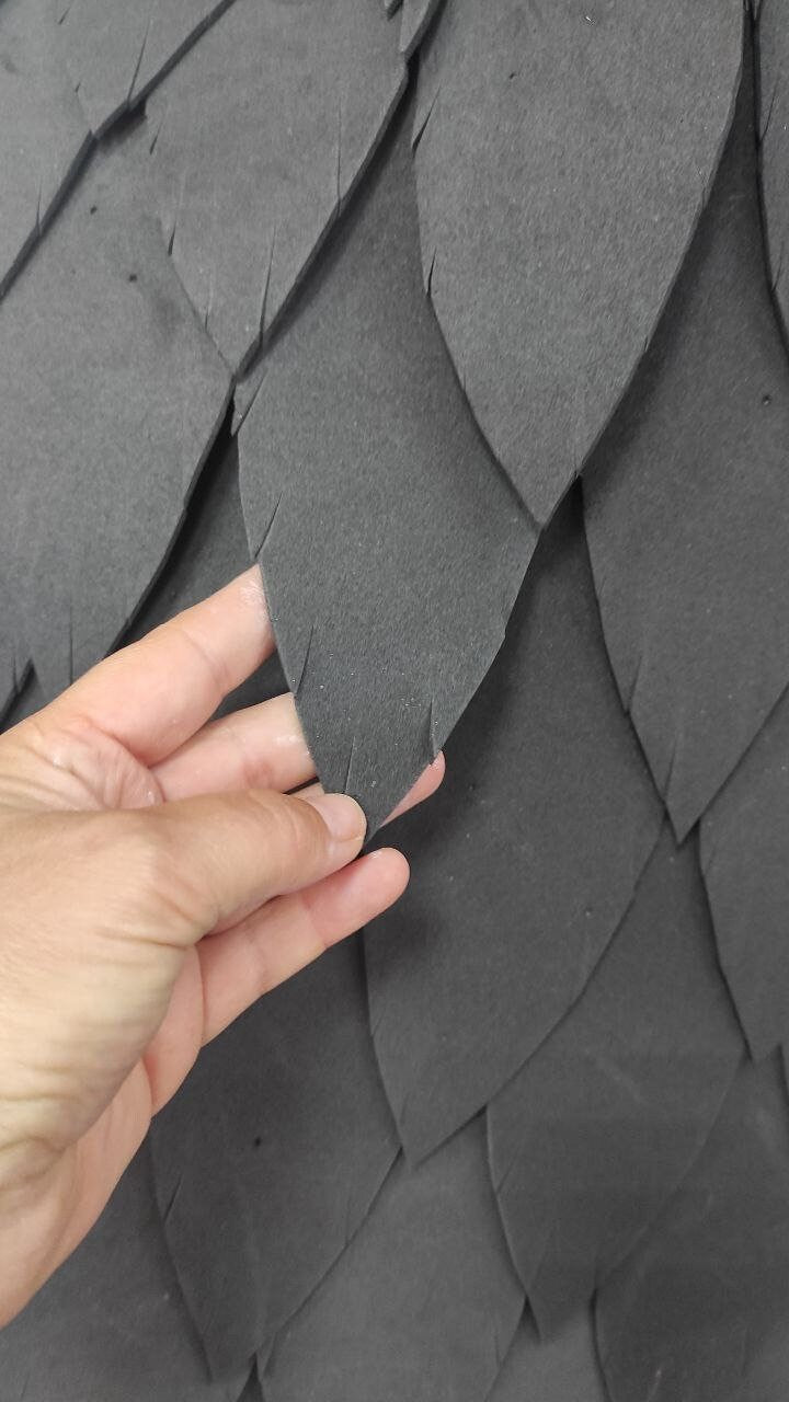 May include: Close-up of a dark gray foam material with a textured, overlapping, leaf-like pattern. The material is soft and flexible, and the leaves are arranged in a staggered pattern.