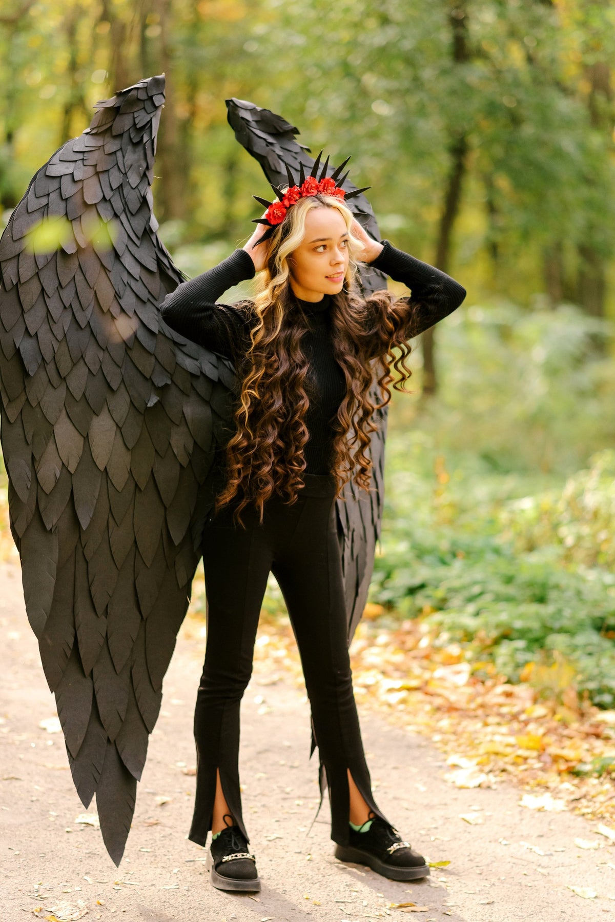 May include: A young woman wearing a black outfit with a crown of red flowers and black feathered wings. She is standing in a forest setting.