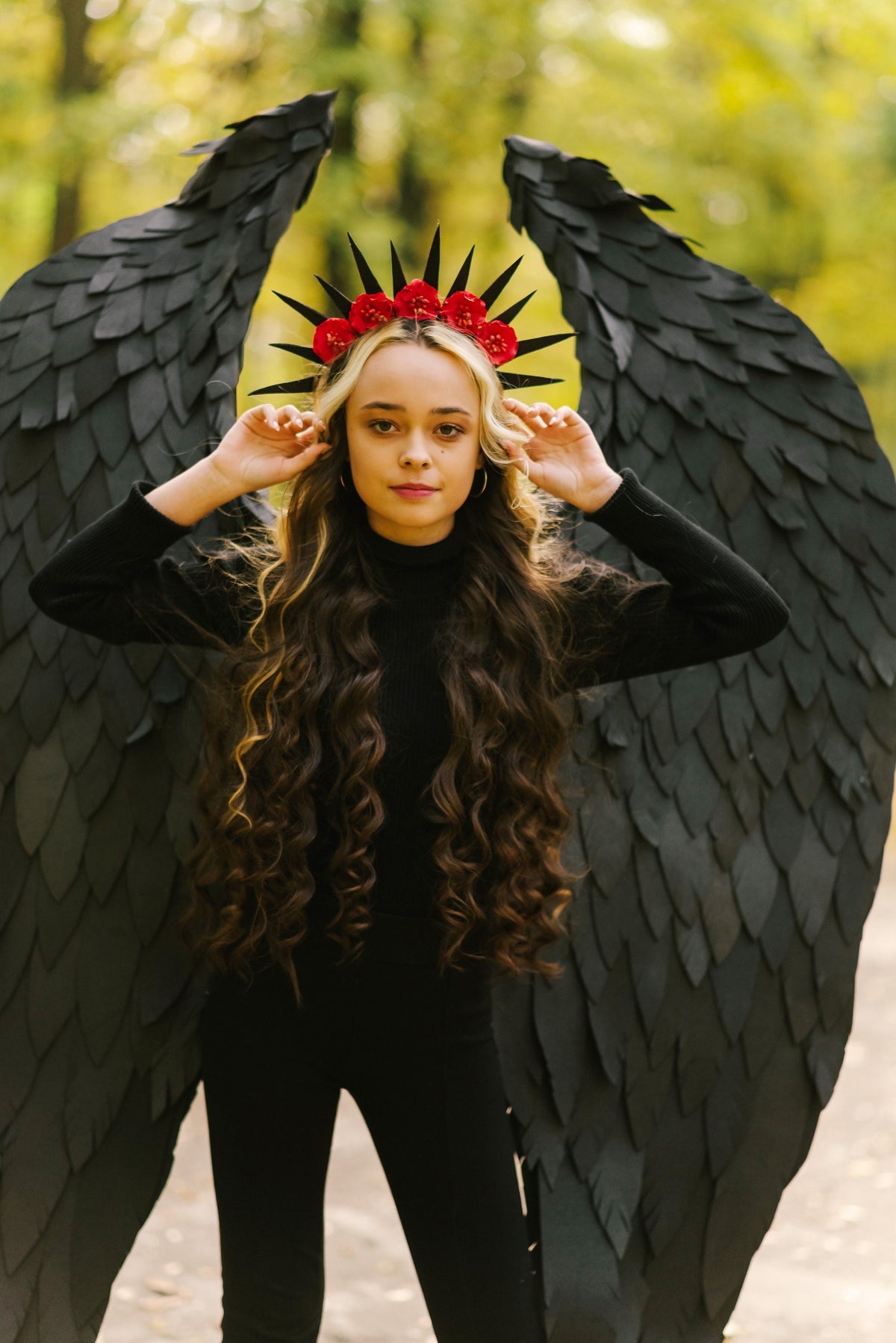 May include: A young person wearing a black outfit with large black wings and a crown of red flowers. The person is standing in a forest setting.