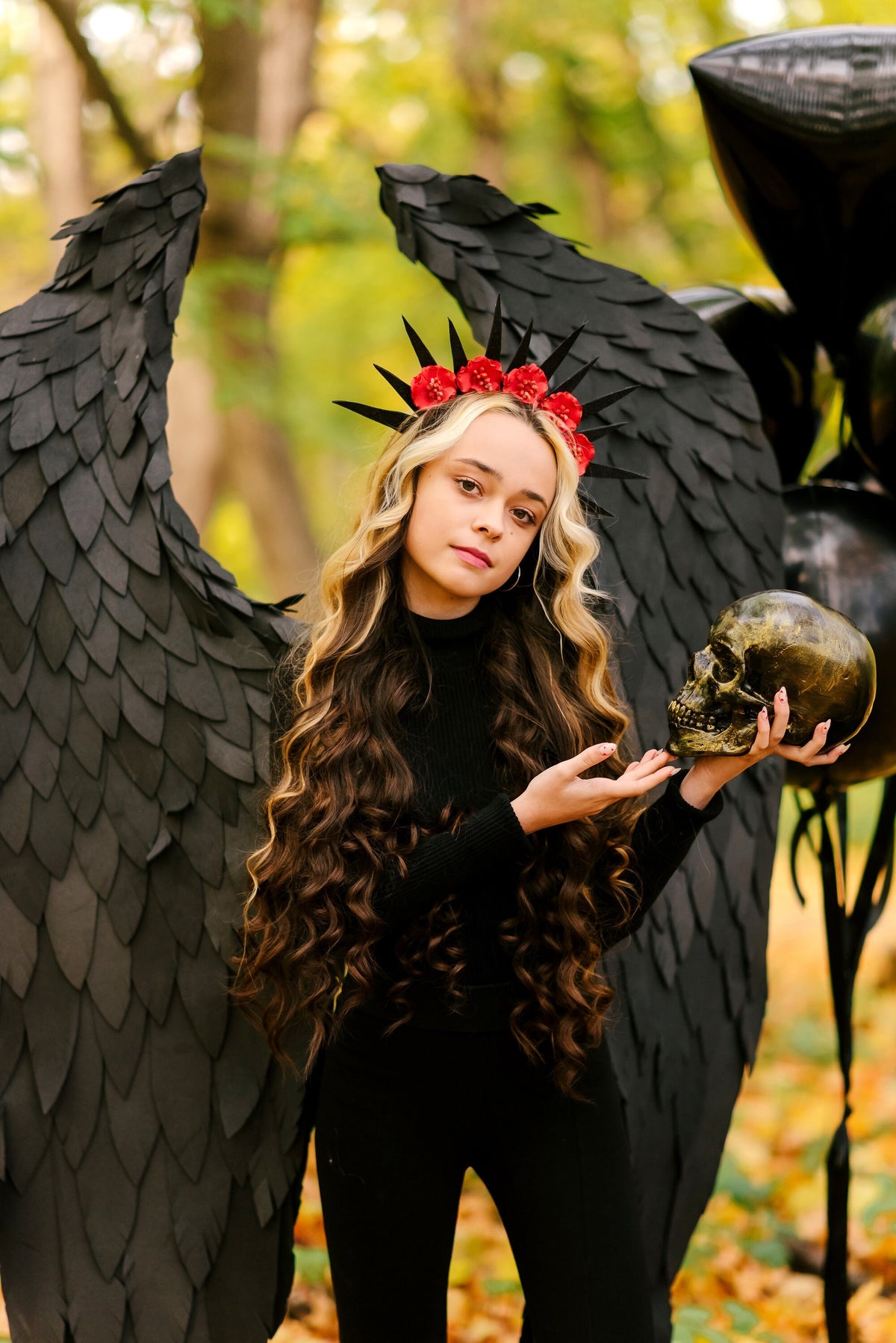 May include: A young person wearing a black outfit with large black wings and a red flower crown, holding a gold skull. The person is standing in a forest setting with fallen leaves on the ground.