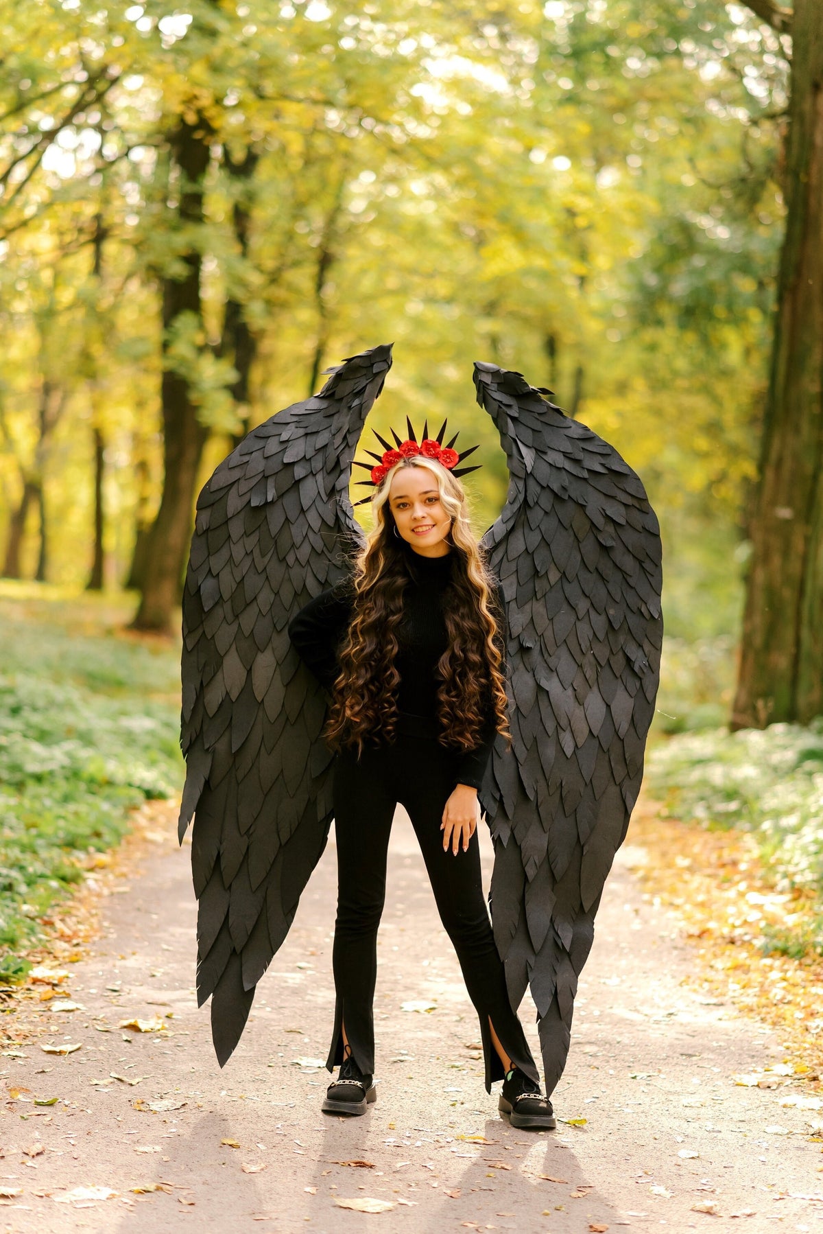 May include: A young woman wearing a black outfit with large black wings and a red flower crown. She is standing on a path in a forest.