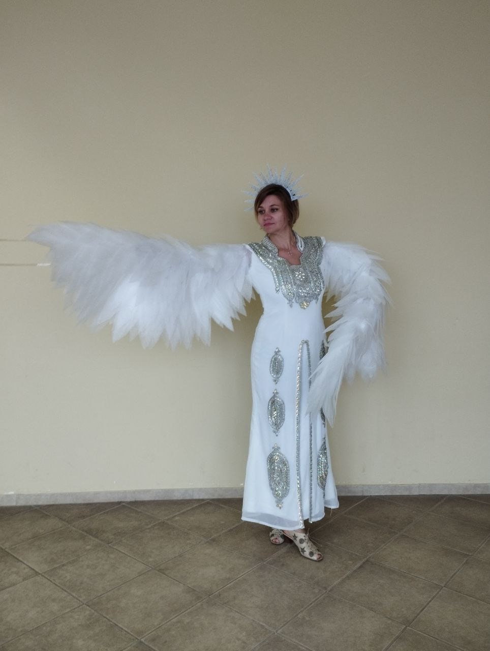 May include: A woman wearing a white gown with silver embellishments and a white feathered angel wing costume. She is wearing a silver crown and is standing on a tiled floor.