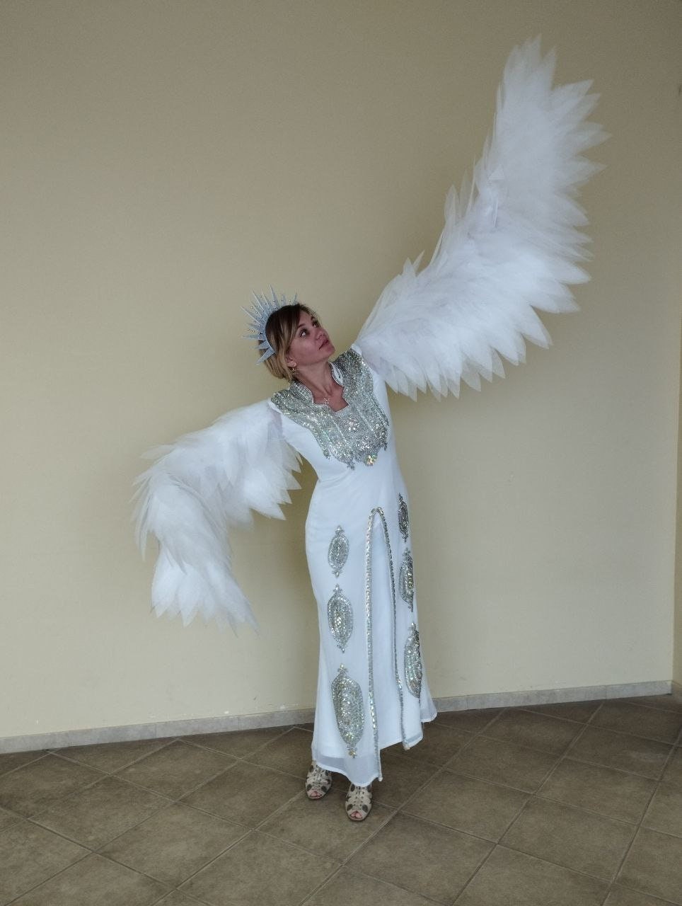 May include: A woman wearing a white gown with silver embellishments and a silver crown, with large white feathered wings outstretched.