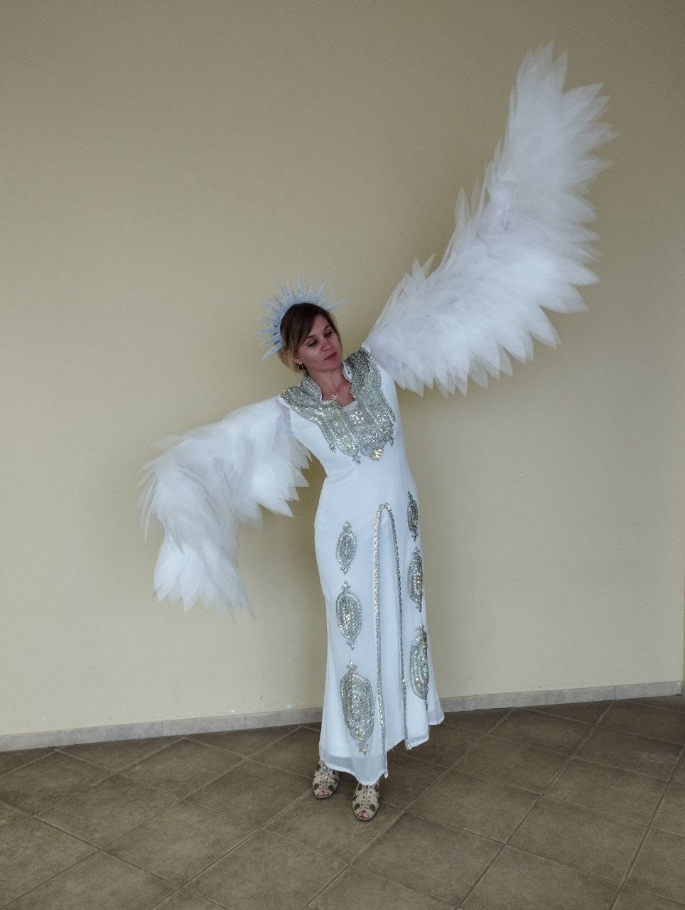 May include: A woman wearing a white gown with silver embellishments and a white feathered angel wing costume. She is wearing a silver crown and has her arms outstretched.