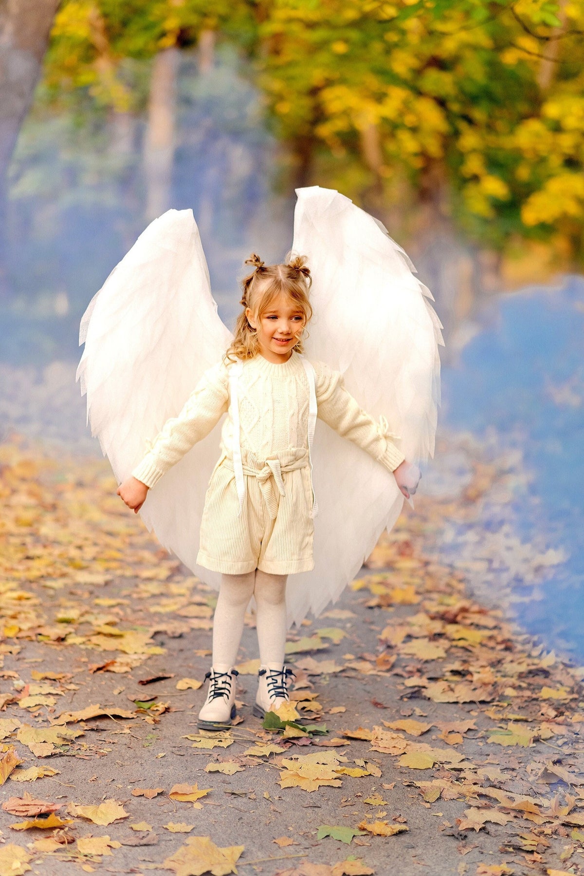 May include: A young girl wearing a white dress and angel wings stands in a park with fallen leaves on the ground. She has her arms outstretched and is looking to the side.