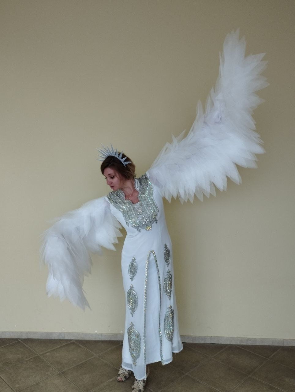 May include: A woman wearing a white gown with silver embellishments and a silver crown. She is wearing large white feathered wings.