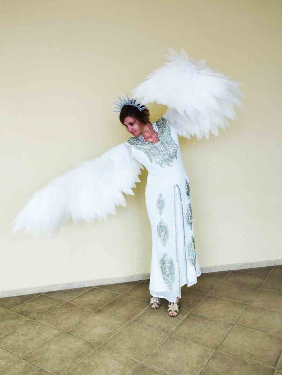 May include: A woman wearing a white dress with silver embellishments and a silver crown, holding large white feathered wings.