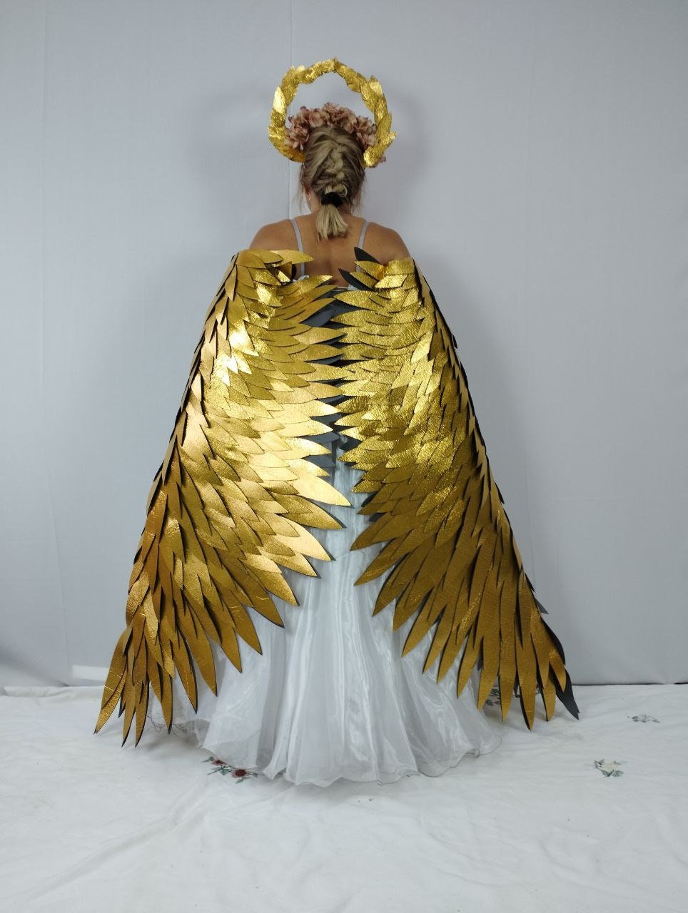 May include: A golden angel wing costume with a matching halo. The wings are large and detailed, with layered golden feathers. The halo is a golden wreath with pink flowers. The costume is worn over a white dress.