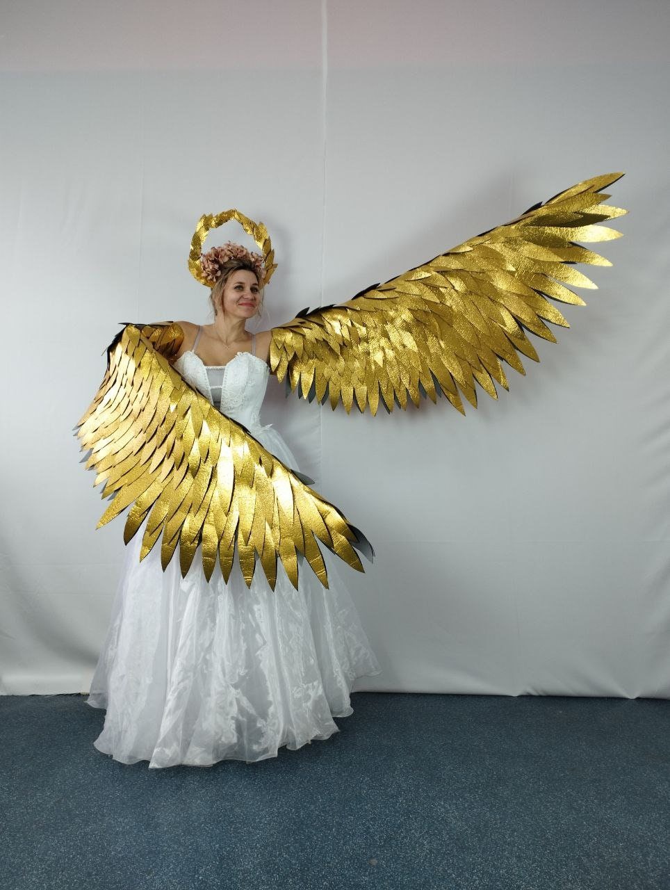 May include: A woman wearing a white dress, gold halo, and large gold angel wings. The wings are made of overlapping, textured gold material. The halo is a gold wreath with pink flowers. The dress is white with a layered skirt.