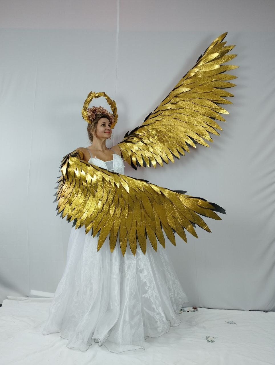 May include: A woman wearing a white dress and large, golden angel wings. The wings are made of layered, textured material, and the woman is also wearing a floral halo. The wings span wide, creating a dramatic effect.