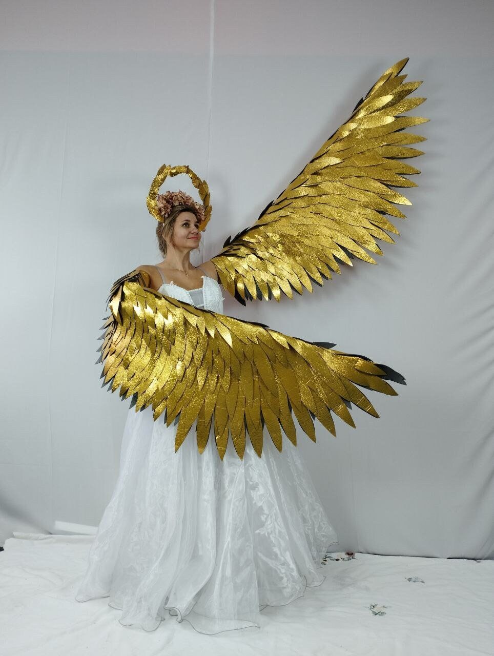 May include: A woman wearing a white dress and a gold halo, with large, gold angel wings. The wings are made of layered, textured material, and the dress has a flowing skirt. The halo is adorned with flowers.