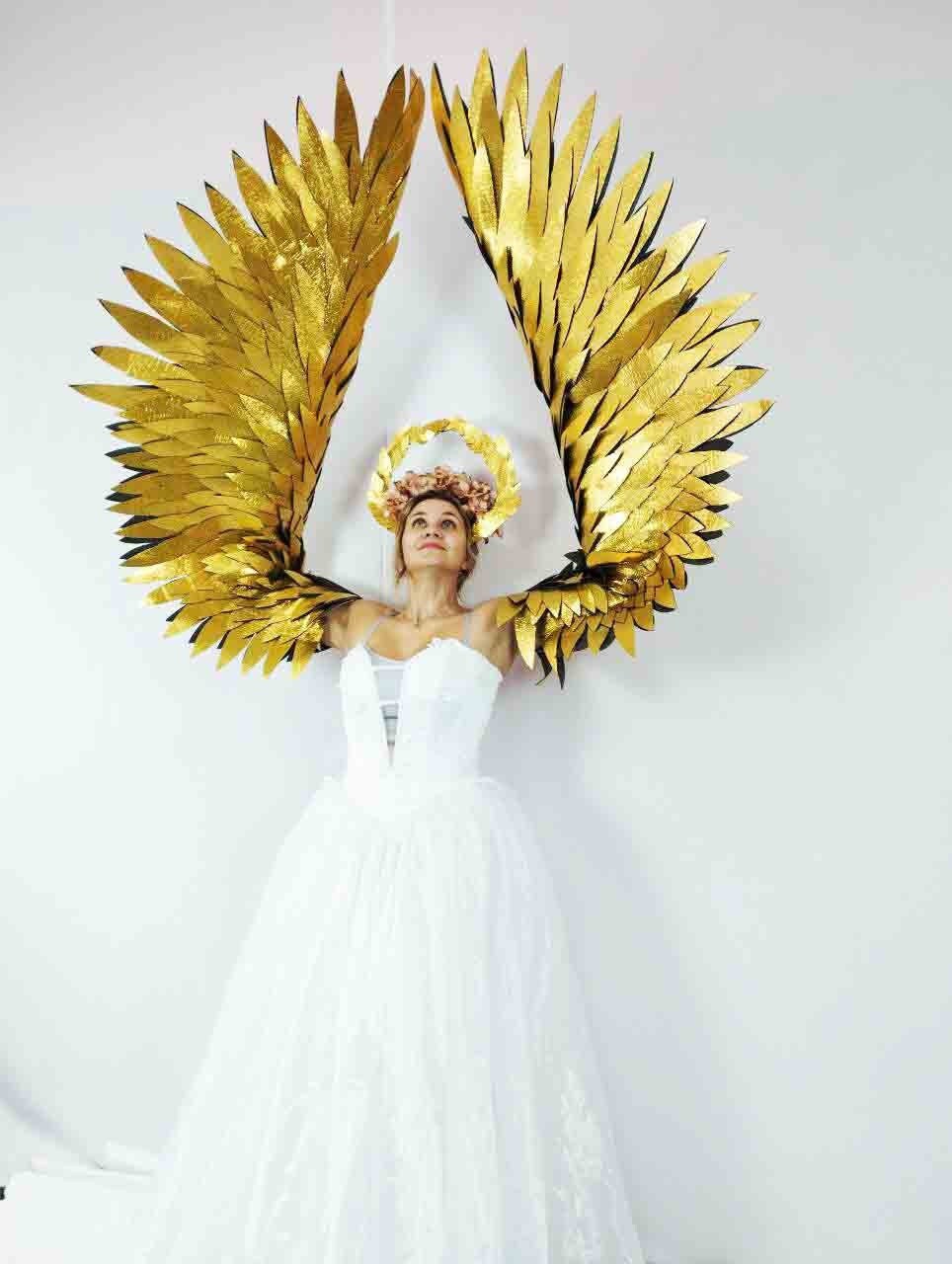 May include: A woman in a white dress with large, golden angel wings. She wears a floral crown. The wings are made of layered, textured gold material, and are spread wide against a white background.