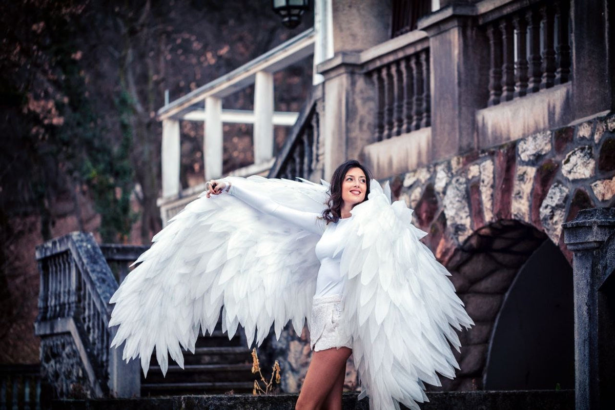 May include: A woman wearing a white dress and large white angel wings stands on a set of stone steps in front of a building.