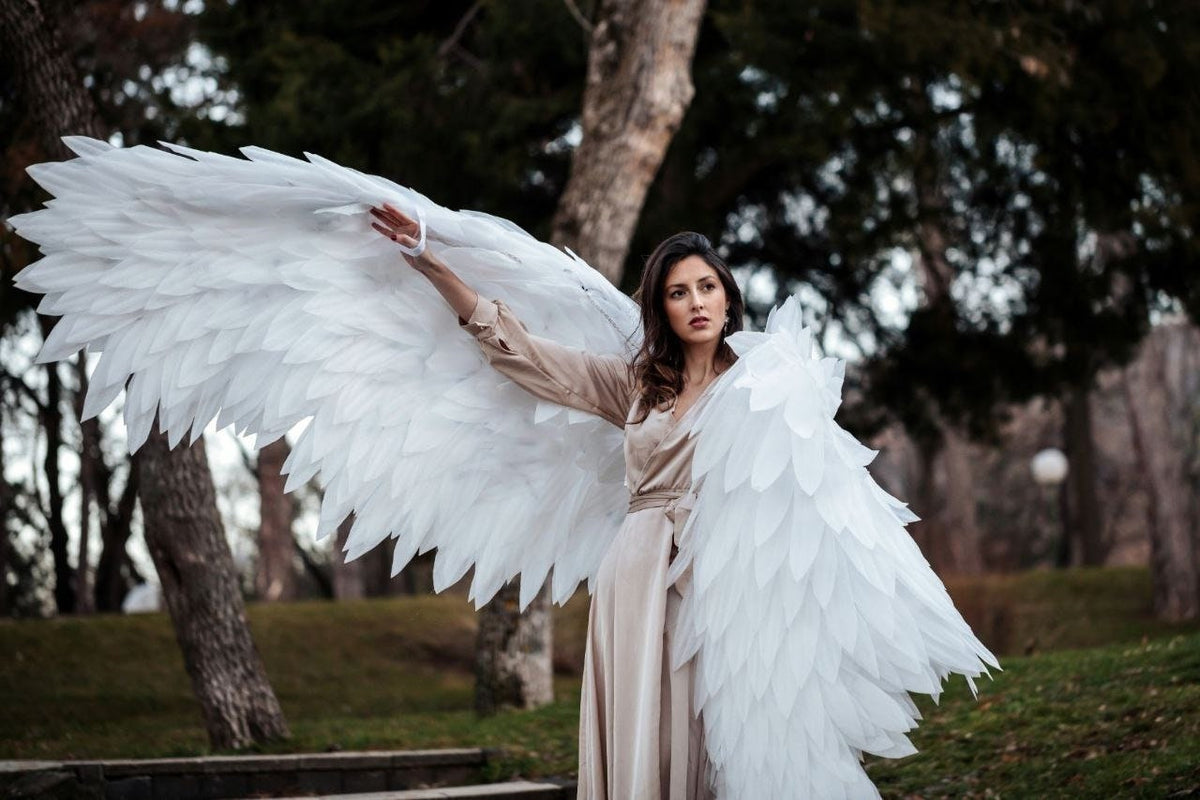 May include: A woman wearing a beige dress with large white angel wings. The wings are made of feathers and are spread out behind her. She is standing in a forest setting.