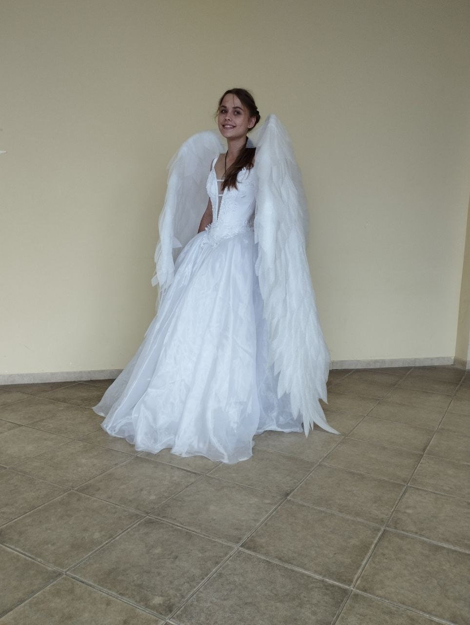 May include: A woman wearing a white dress with large white angel wings. She is standing on a tiled floor.