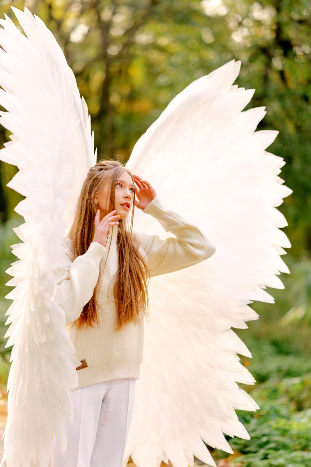 May include: A young person with long blonde hair wearing a white sweater and white pants, standing in a forest with large white angel wings.