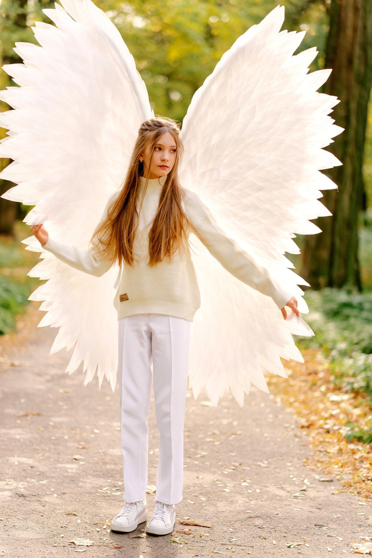 May include: A young person wearing a white sweater and white pants with large white angel wings. The person is standing on a path in a forest with fallen leaves.
