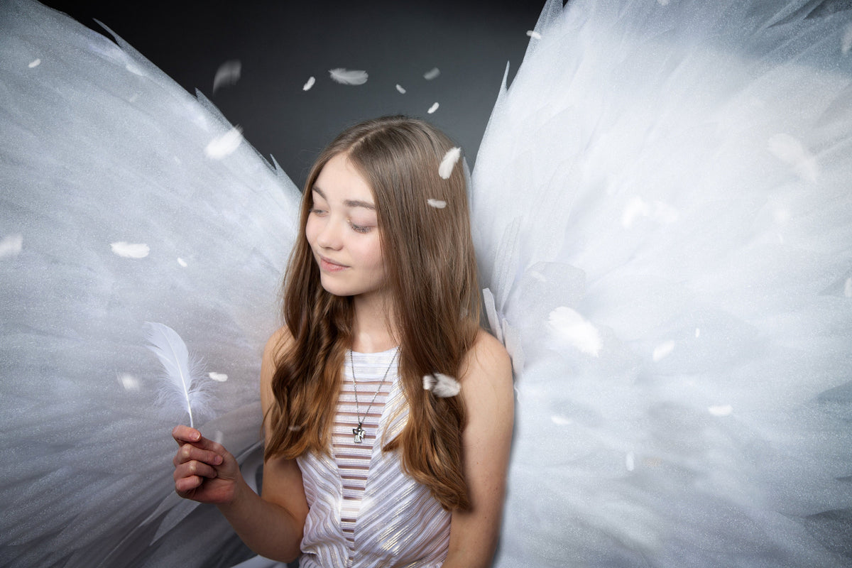 May include: A young person with long brown hair wearing a white dress with a silver necklace and holding a white feather. The person is standing in front of a white background with white feathers falling around them.