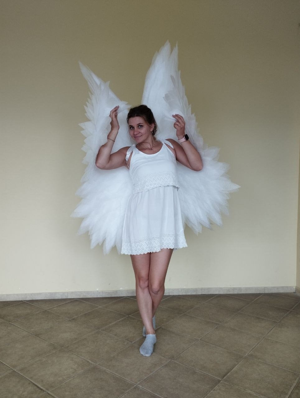May include: A woman wearing a white dress and white angel wings. She is standing on a tiled floor.