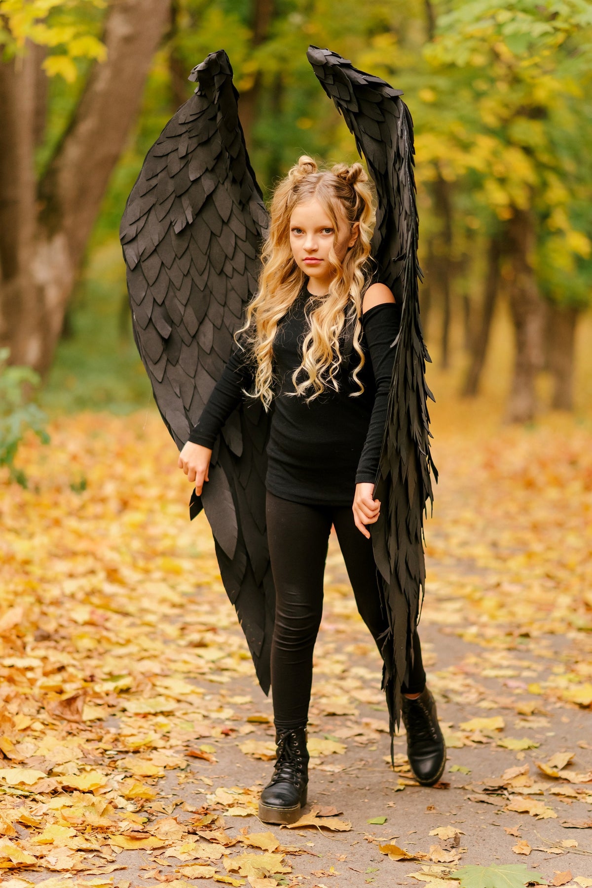May include: A young person wearing a black costume with large black wings, standing on a path covered in fallen autumn leaves.