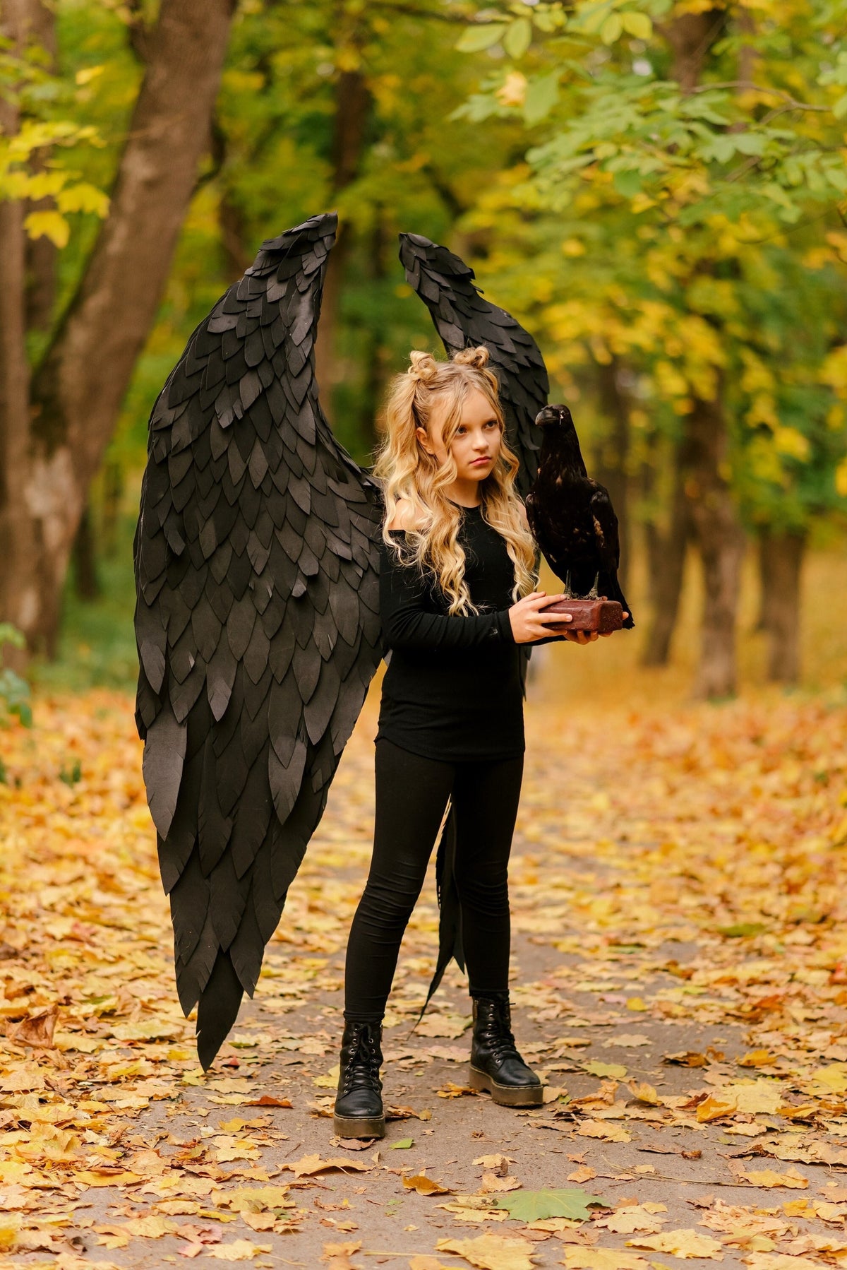 May include: A young person wearing a black costume with large black wings and holding a black raven. The person is standing on a path covered in fallen autumn leaves.