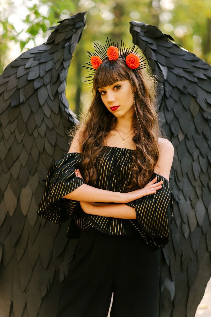 May include: A woman wearing a black and gold striped off-the-shoulder top and black pants. She is wearing a black headband with red flowers and has black wings behind her.