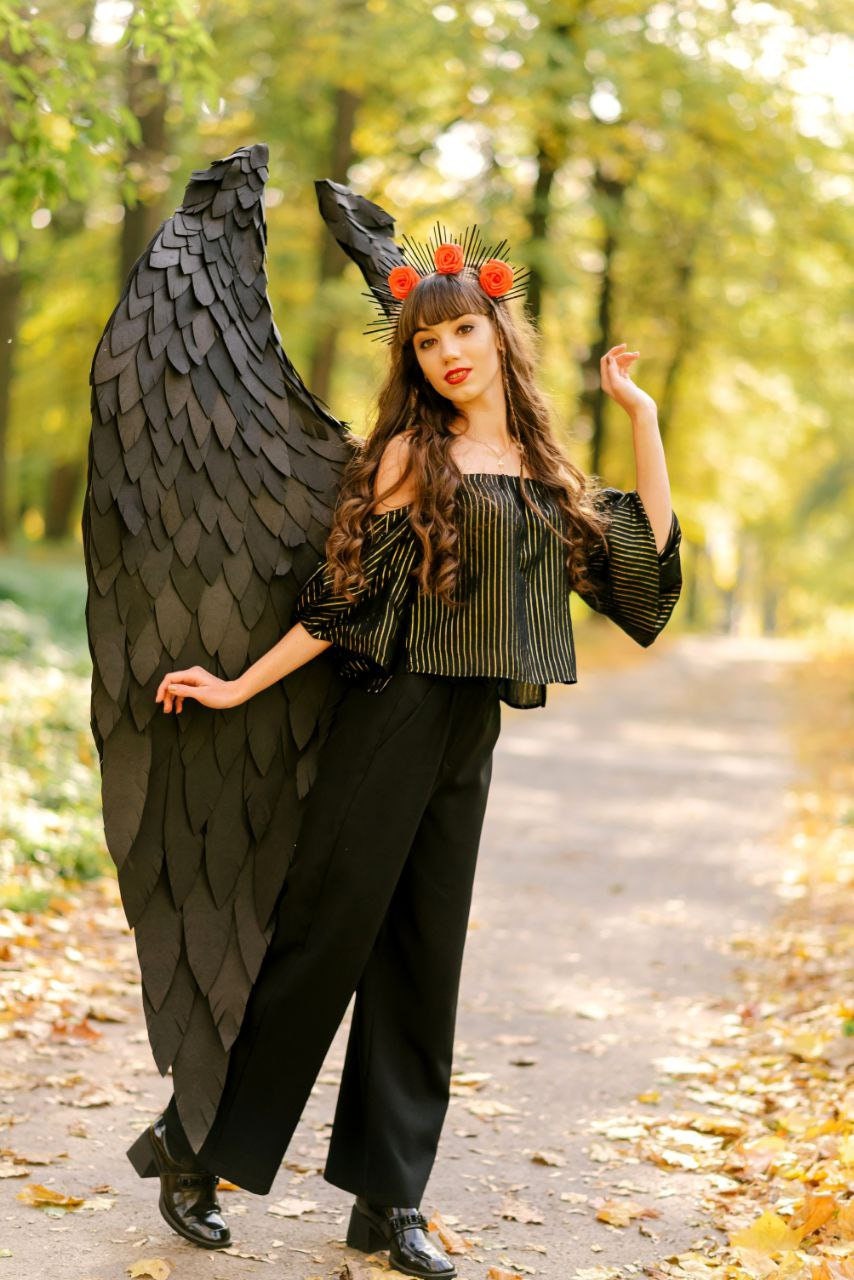 May include: A woman wearing a black and gold striped top and black pants with a black feathered angel wing and a red flower crown.