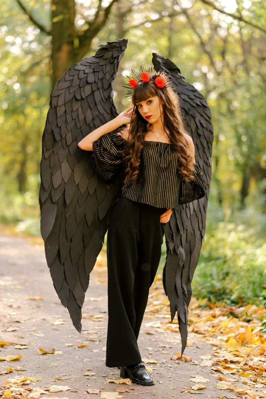 May include: A woman wearing a black and gold striped top and black pants with large black angel wings. She is wearing a crown of red flowers on her head.