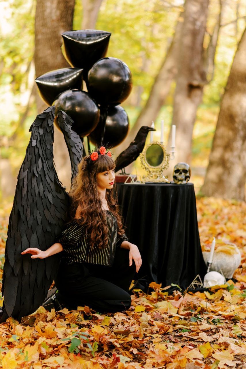 May include: A young person dressed in black with black feathered wings and a red flower crown sits in a forest setting.  A black raven, a skull, and a mirror are on a black table with candles.  Black balloons are in the background.