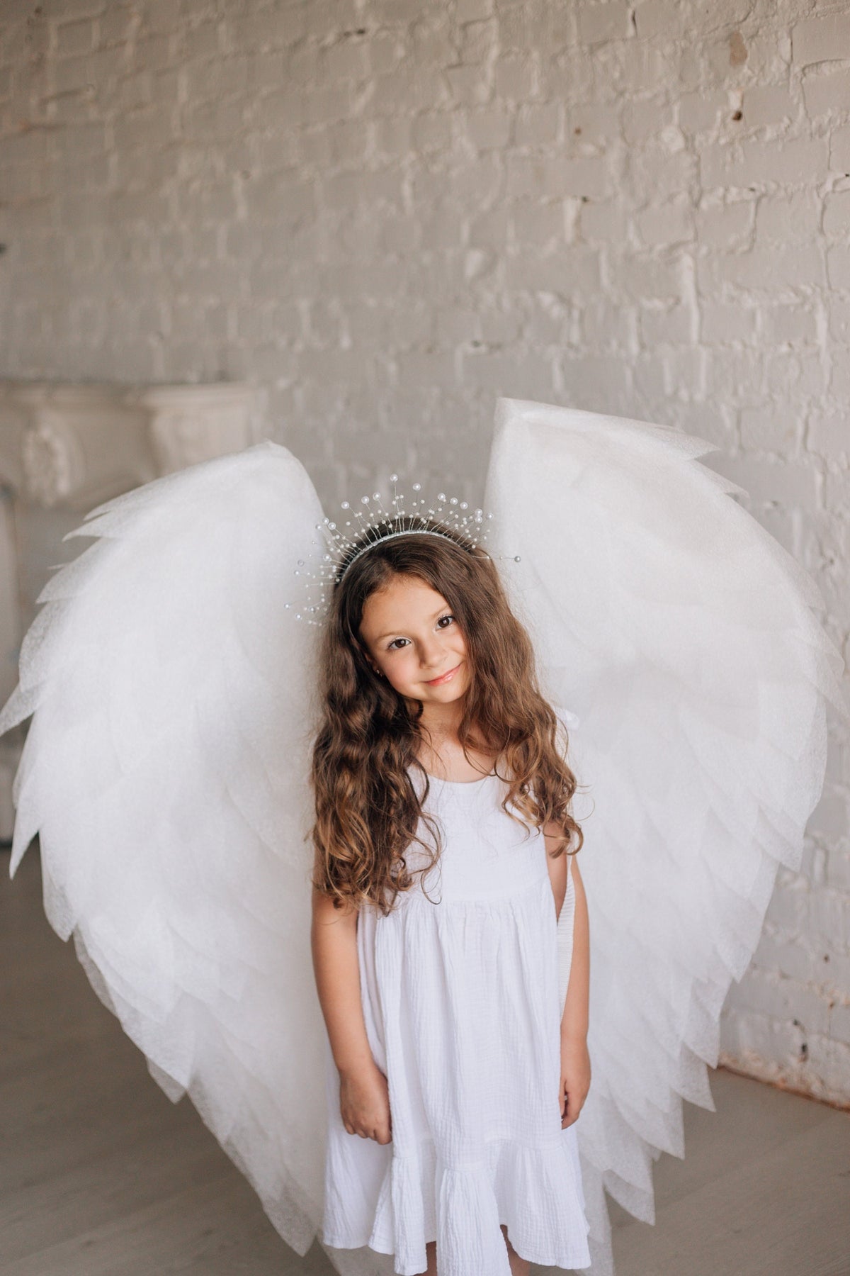 May include: A young person wearing a white dress, a silver beaded crown, and large white angel wings. The wings are layered and detailed. The background is a white brick wall.