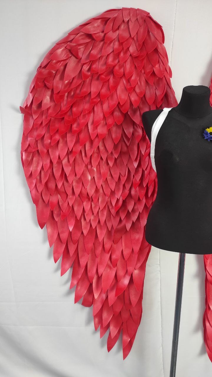 May include: Large, vibrant red angel wings with a layered, feather-like design. The wings are displayed next to a black mannequin torso with a white strap. The wings are a striking costume piece.