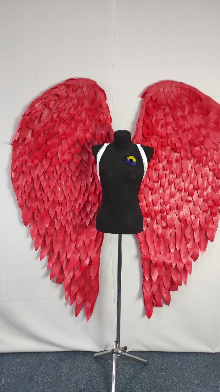 May include: A pair of large, vibrant red angel wings with a layered feather design. A black mannequin is positioned in front of the wings, wearing a white harness and a small blue and yellow flower pin. The wings are a striking decorative piece.