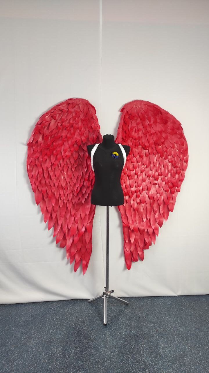 May include: Large, vibrant red angel wings with a detailed feather design. The wings are displayed on a black mannequin with white straps and a small floral pin. The wings are a striking decorative piece.