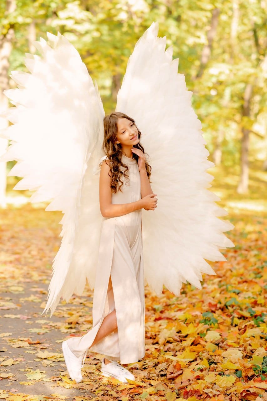 May include: A young person wearing a white dress and large white angel wings stands in a forest setting with fallen autumn leaves on the ground.