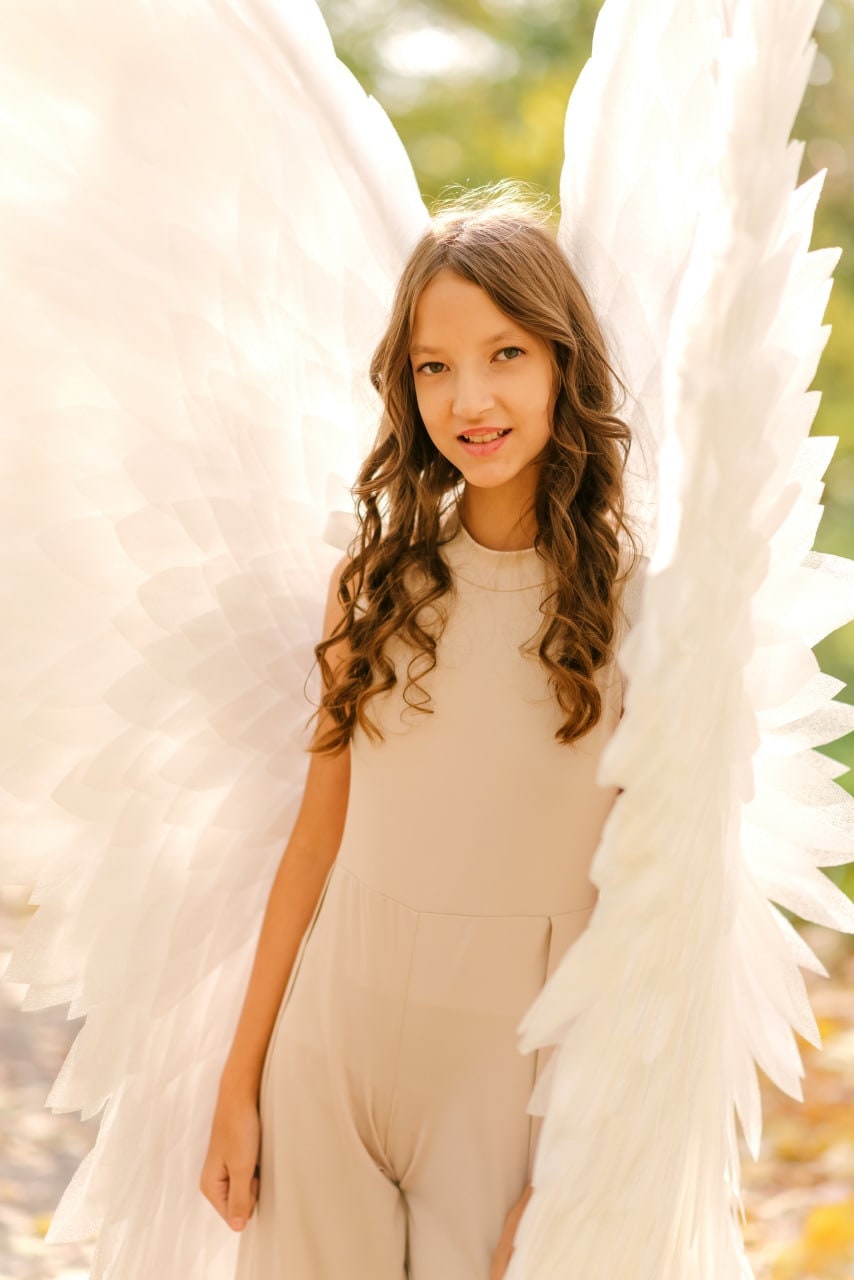 May include: A young person wearing a beige jumpsuit with large white angel wings. The wings are made of feathers and are spread out behind the person.