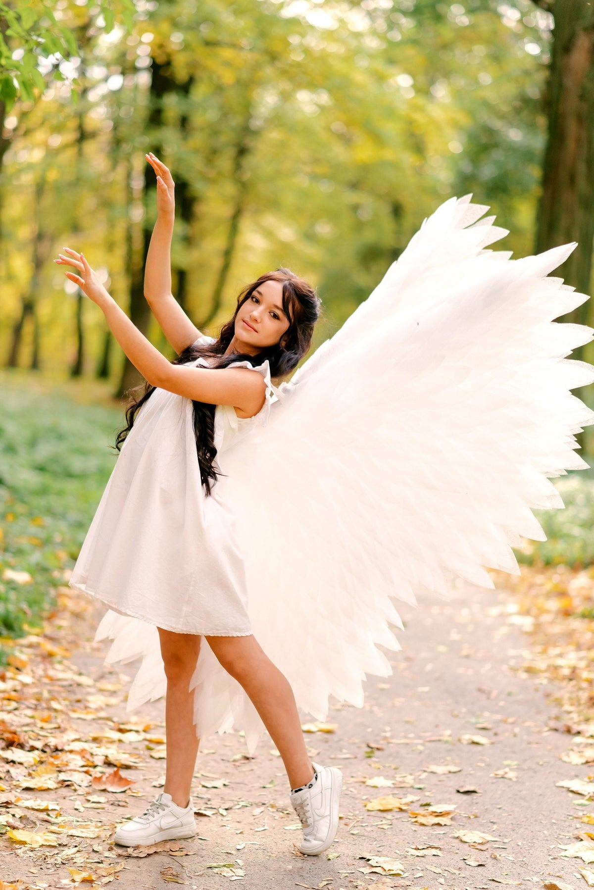 May include: A woman wearing a white dress and white angel wings stands in a forest with fallen leaves. She is looking at the camera and smiling.