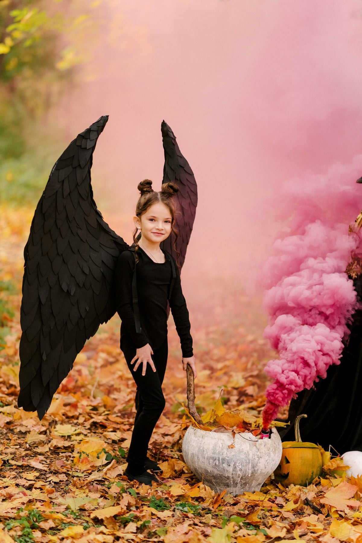 May include: A young girl dressed in black with black angel wings stands in a forest setting with pink smoke behind her. She is holding a wooden stick and a cauldron is in front of her.