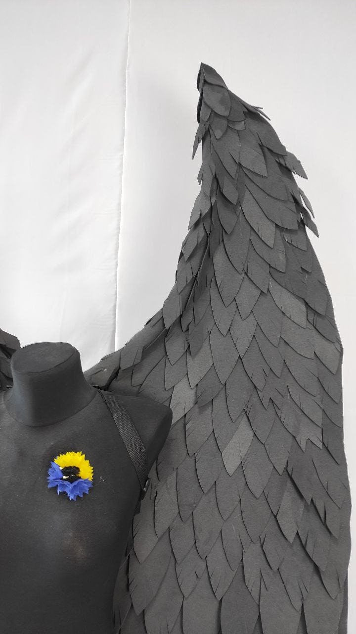 May include: A pair of black angel wings made of paper feathers. The wings are attached to a black mannequin with a blue and yellow flower pin.