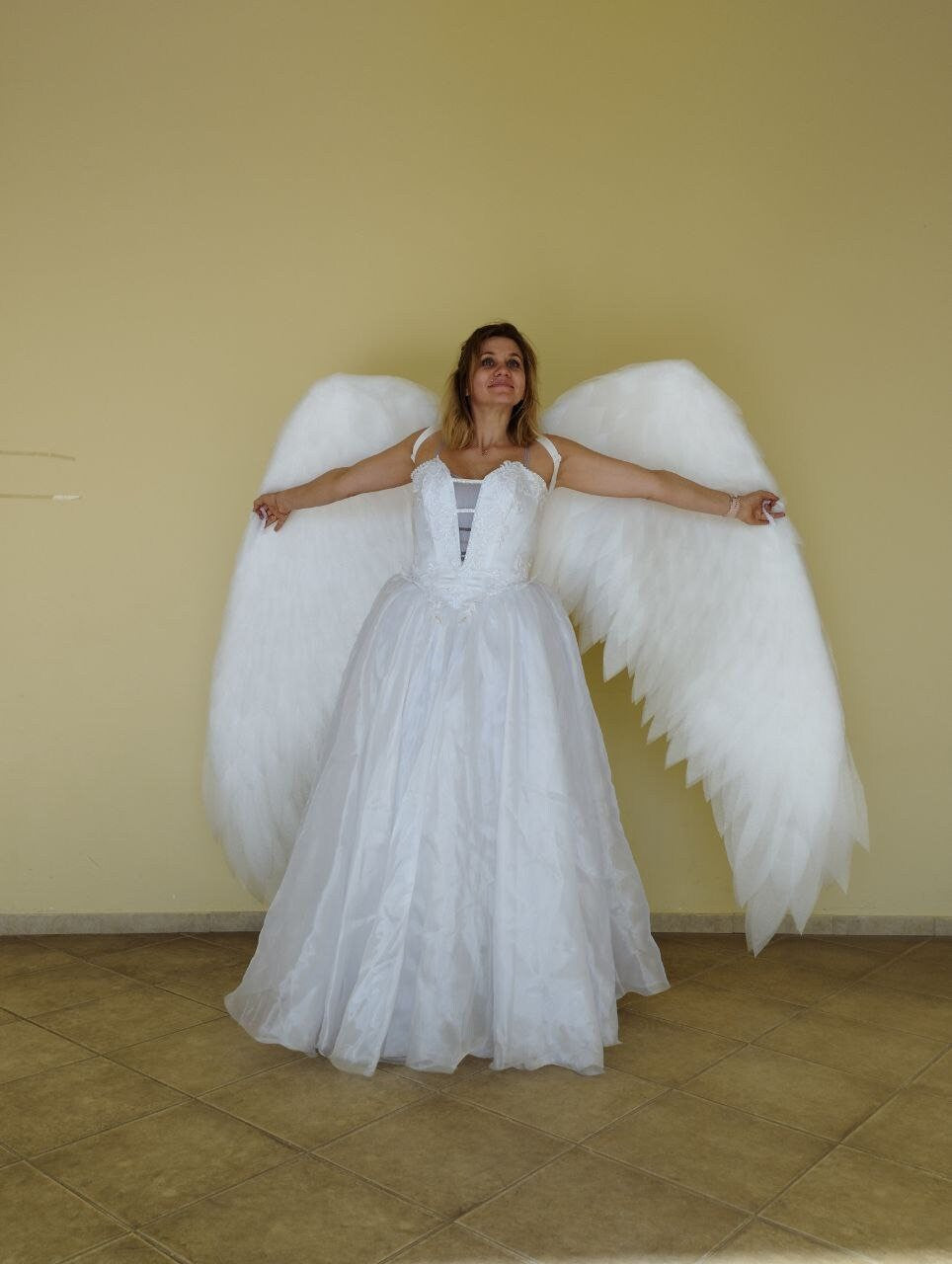May include: A white wedding dress with a bodice and a flowing skirt. Large, fluffy white angel wings are attached to the back. The person is standing with arms outstretched.