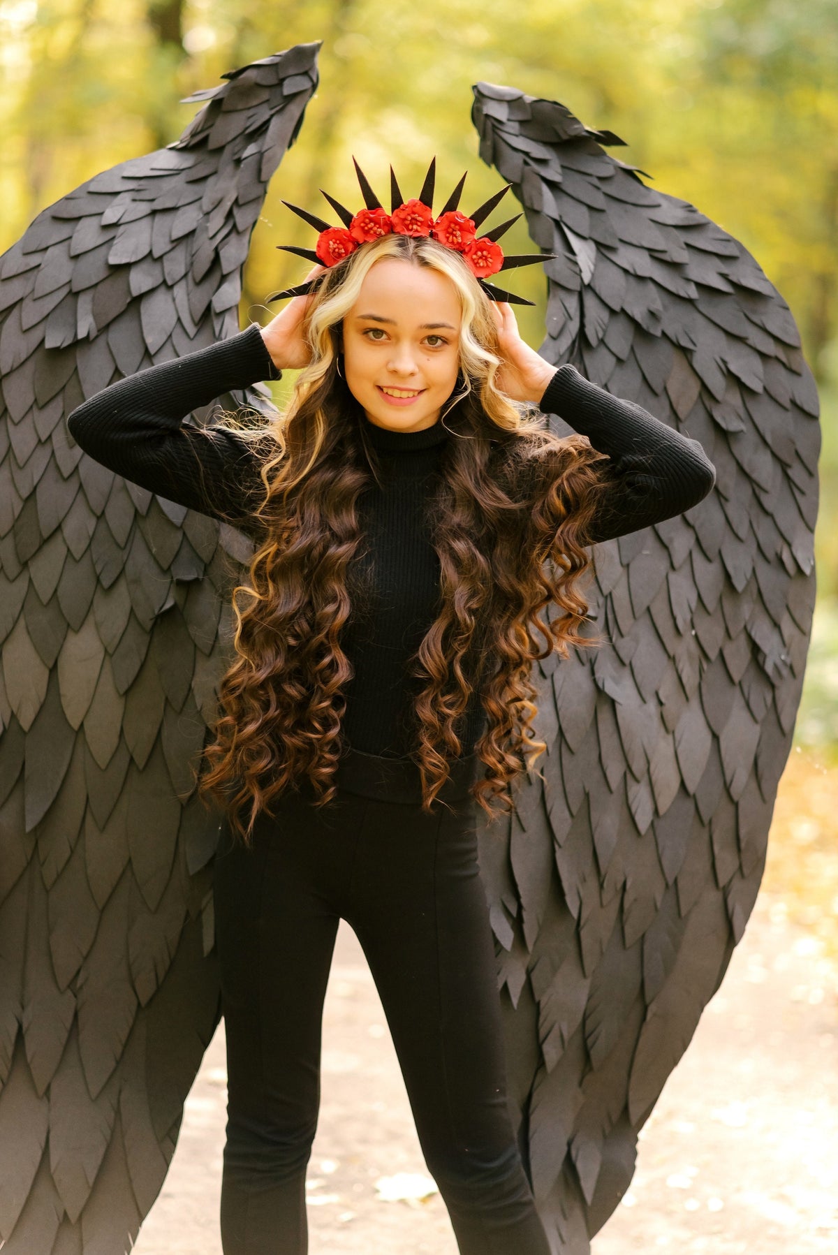 May include: A young woman with long brown hair wearing a black turtleneck shirt and black pants. She is wearing a crown of red flowers and black wings.