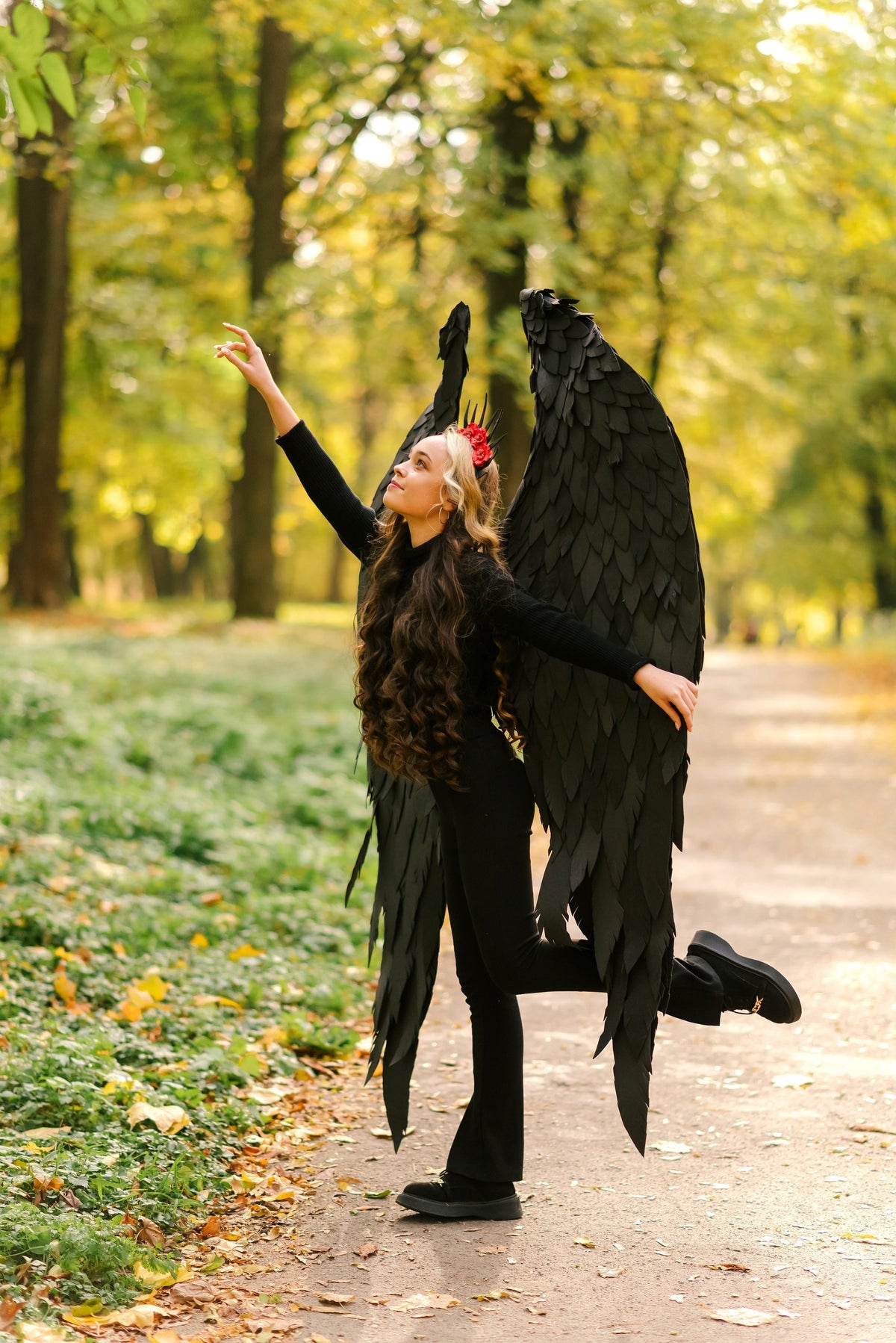 May include: A young person wearing a black outfit with large black feathered wings and a red flower crown, standing in a forest setting.
