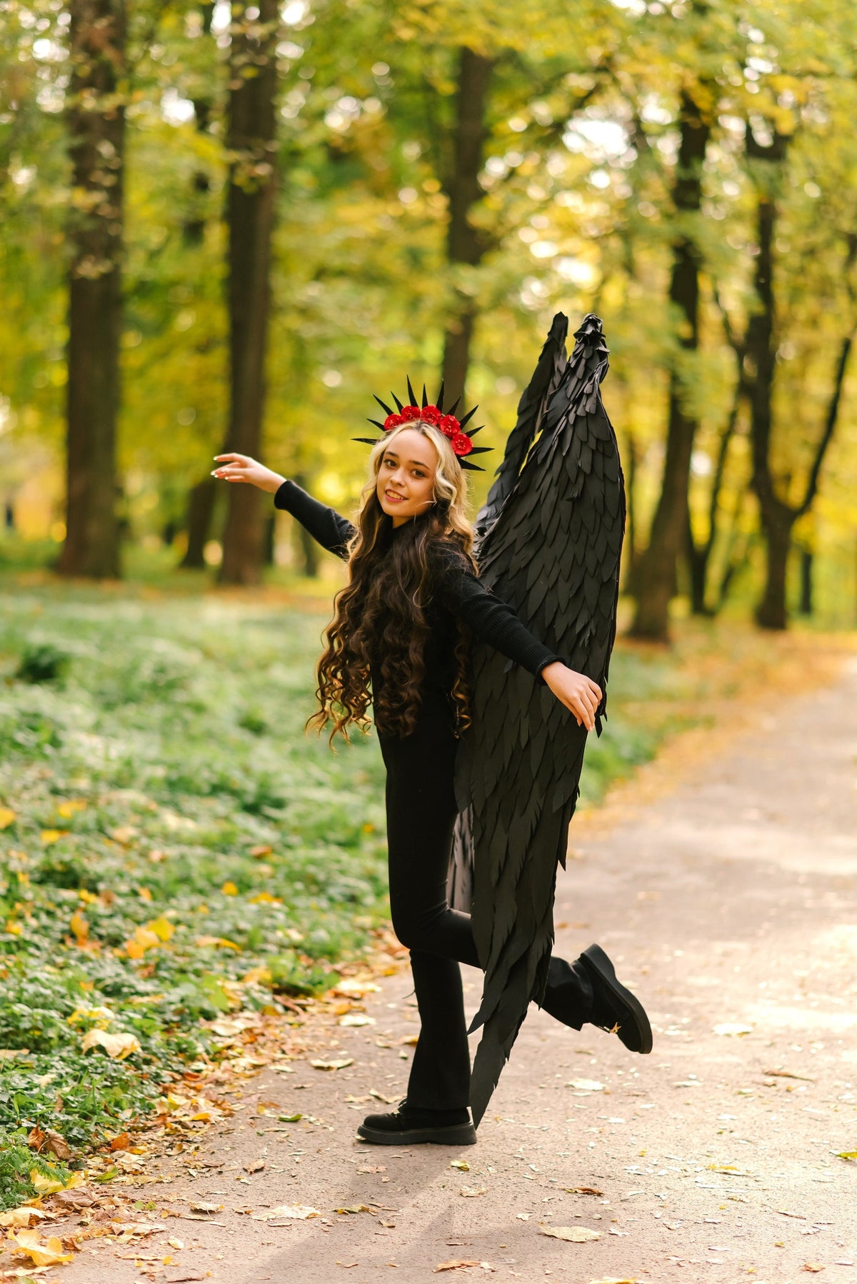 May include: A young person wearing a black outfit with large black wings and a red flower crown. They are standing on a path in a park with fall foliage.