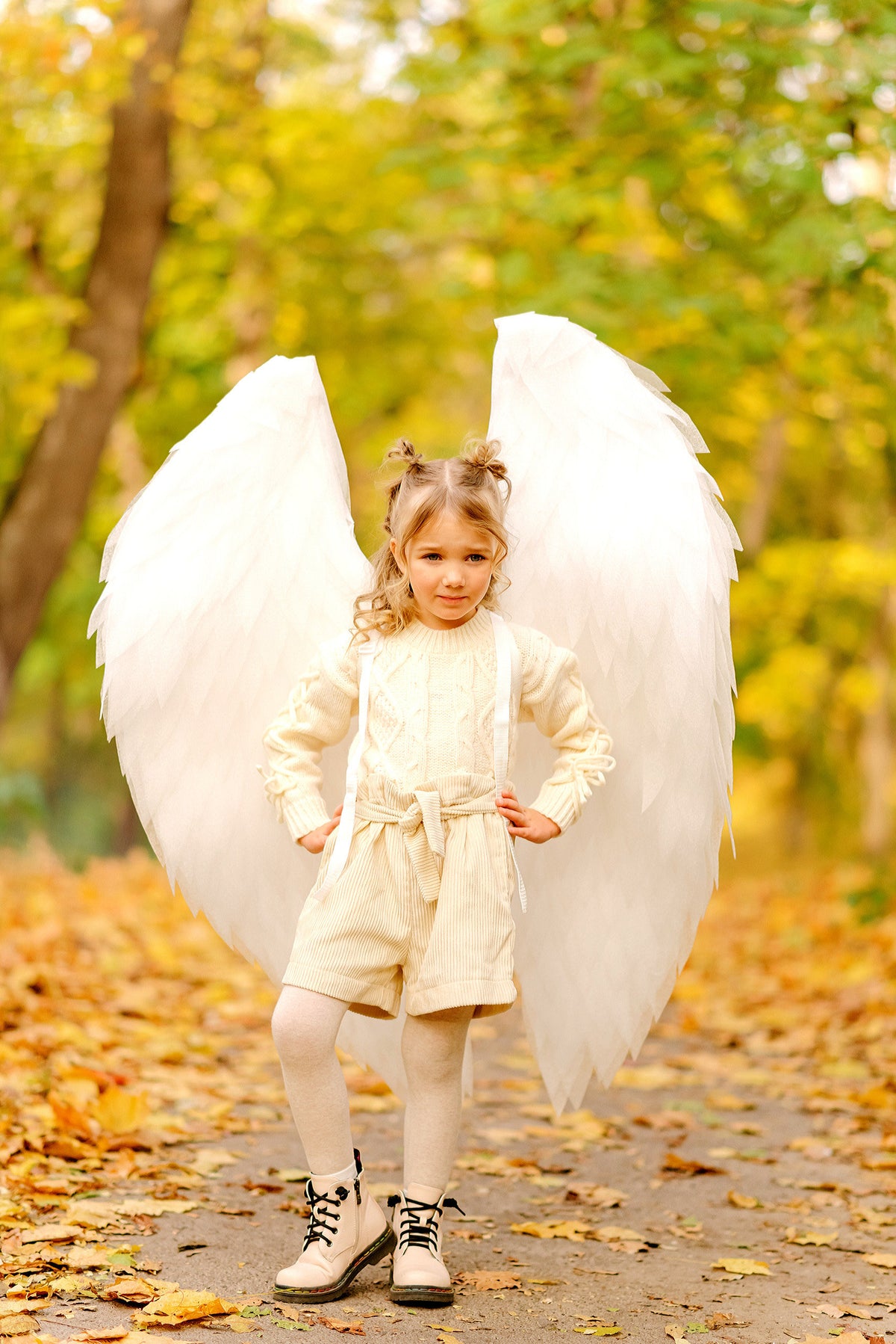 May include: A young girl wearing a white sweater and white corduroy shorts with a white angel wing costume. She is standing in a park with fallen autumn leaves on the ground.