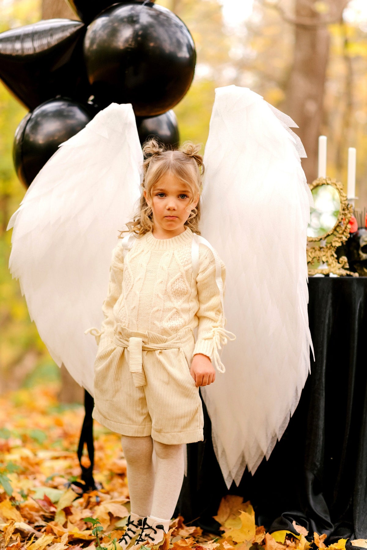 May include: A young girl wearing a white sweater and cream-colored shorts with a white angel wing costume. She is standing in a forest with autumn leaves on the ground.