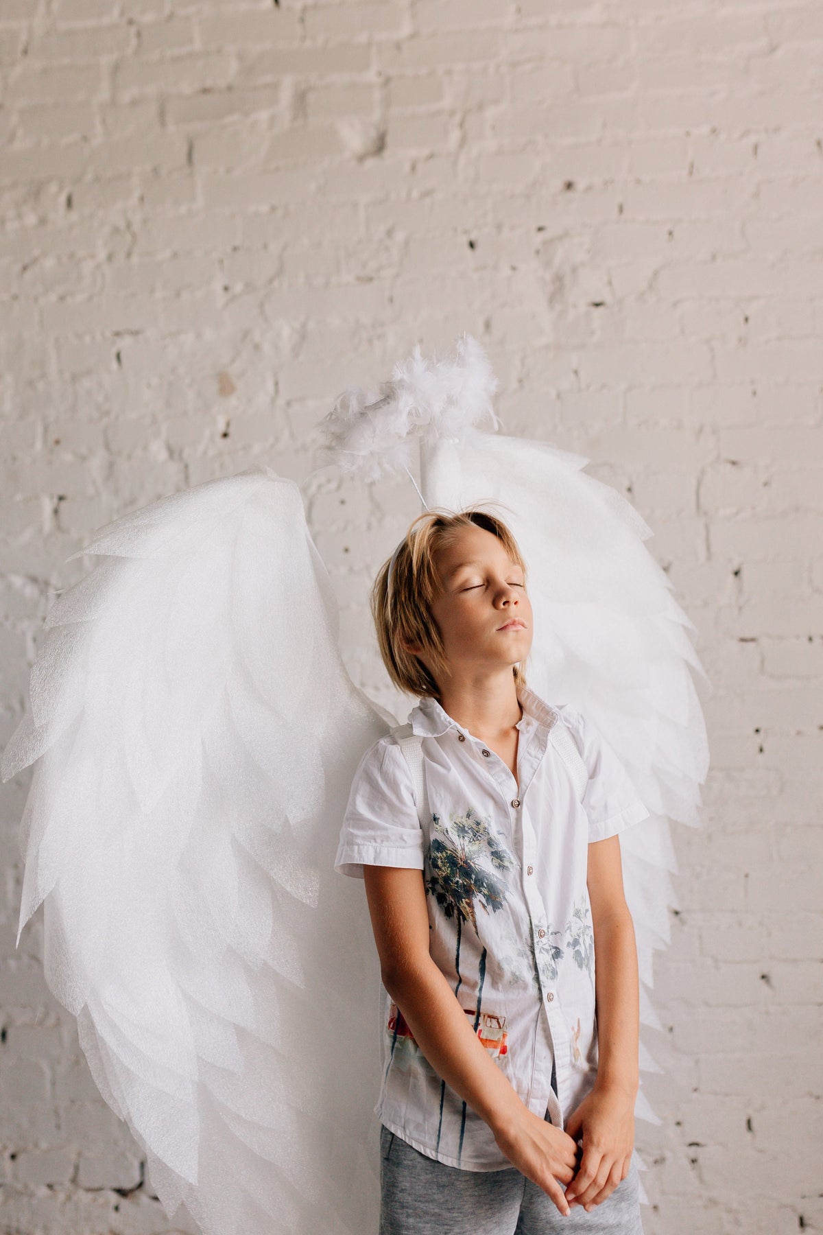 May include: A young person wearing a white shirt with a palm tree print and a white angel costume with wings and a halo. The person has their eyes closed and is standing in front of a white brick wall.