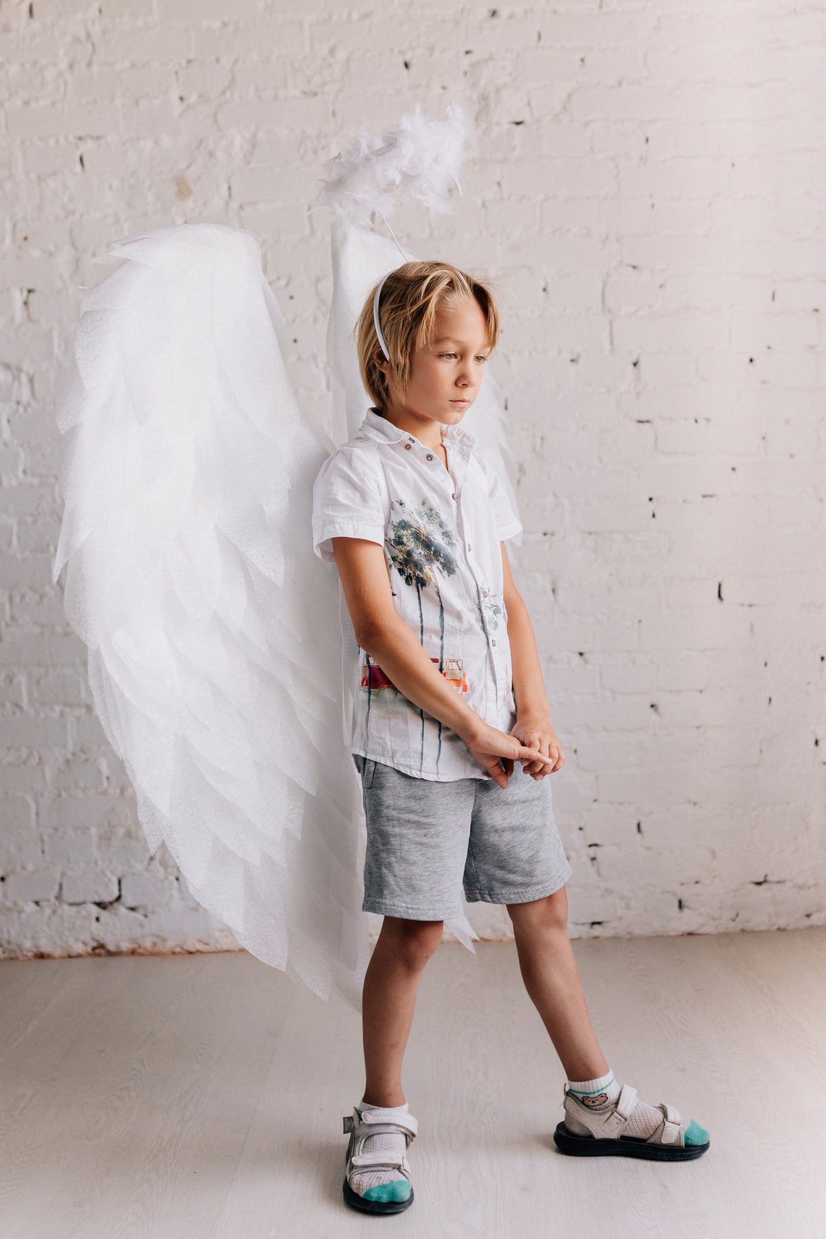 May include: A young boy wearing a white shirt with a palm tree print and gray shorts. He is wearing a white angel wing costume with a halo headband. The boy is standing in front of a white brick wall.