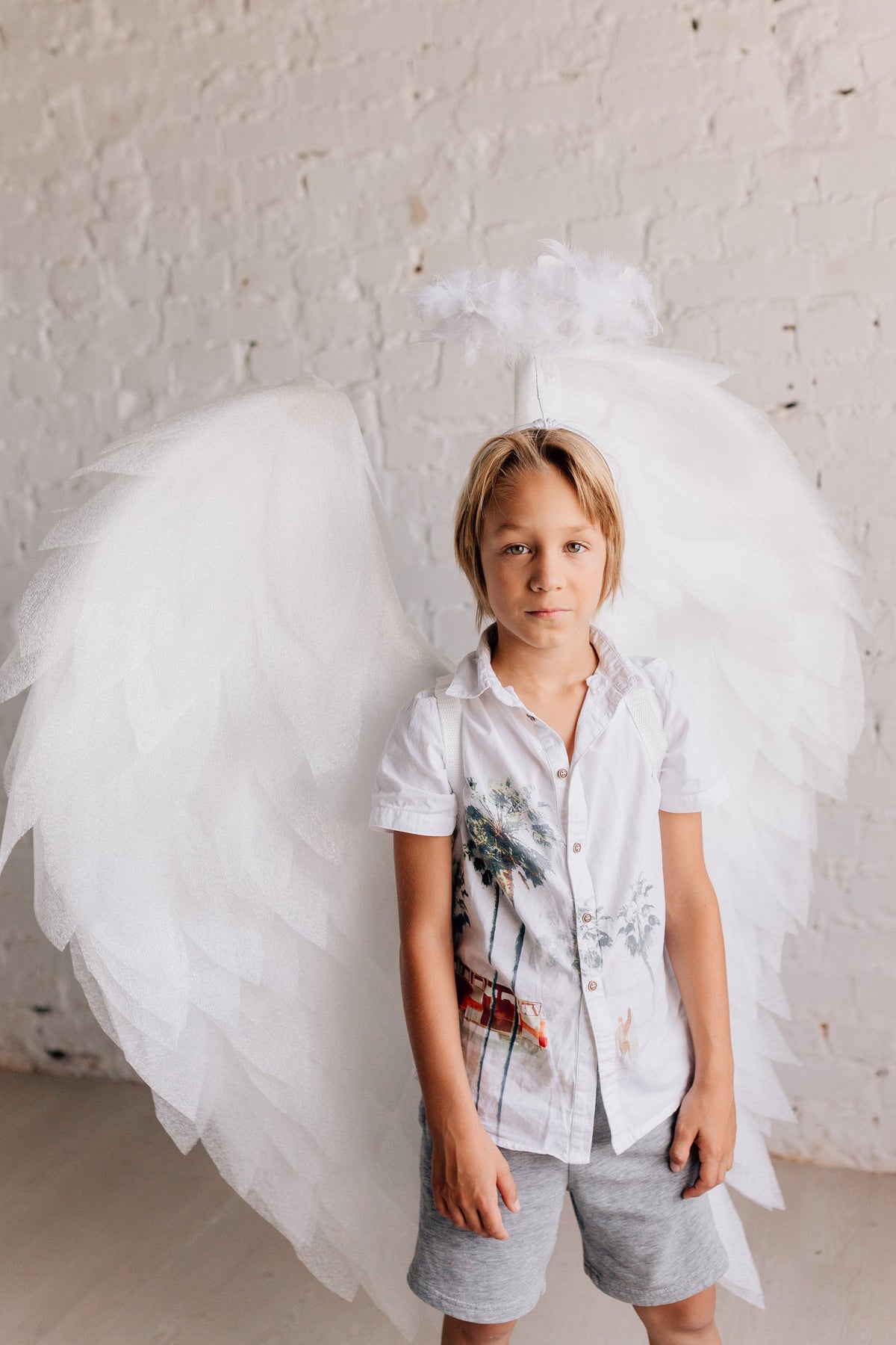 May include: A young person wearing a white shirt with a palm tree print and gray shorts. They are wearing a white angel costume with wings and a halo.