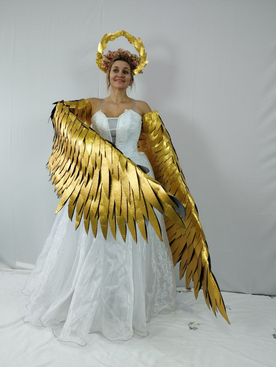 May include: A woman wearing a white dress, gold angel wings, and a gold halo. The wings are large and detailed, with a layered design. The halo is circular with leaf-like details. The dress is white and has a layered skirt.